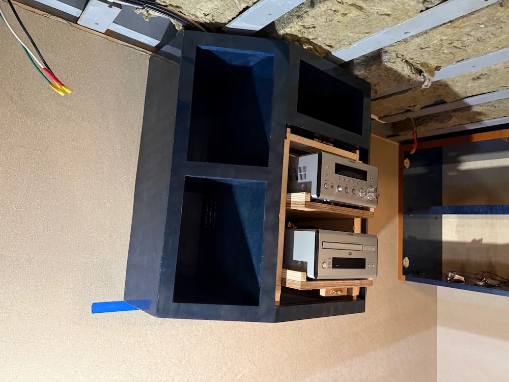 A wooden shelf with electronic equipment including an amplifier and two other devices, placed in a room under construction with exposed insulation and wiring.