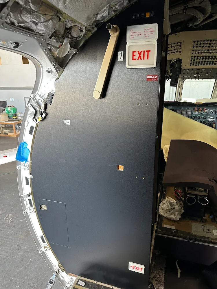 Interior of an aircraft cockpit showing a panel with an 'EXIT' sign, a beige handle, and various control panels and wires.