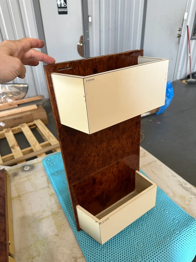 A vintage wooden snack vending machine with two white compartments labeled for different soda cans, placed on a workbench in a workshop.