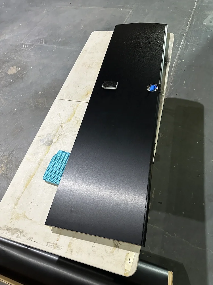 A black safe with a keypad lock and blue dial on top, resting on a white table with a blue textured mat nearby, on a gray concrete floor.