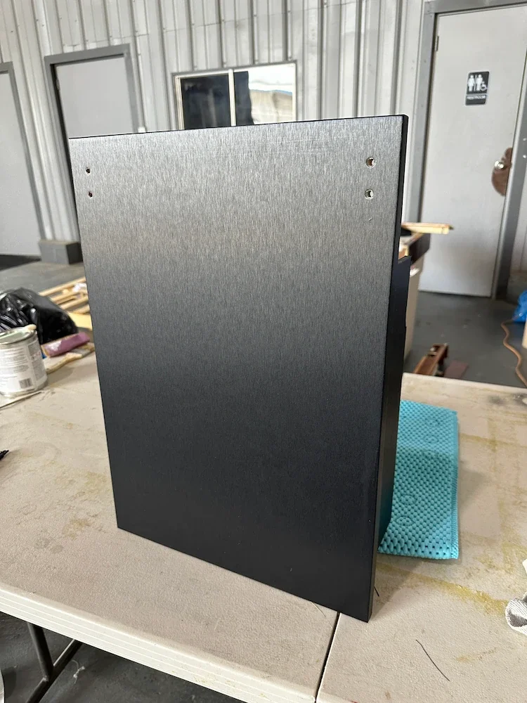 A black, rectangular electronic device, possibly a speaker or small appliance, on a beige work table in a workshop or garage.