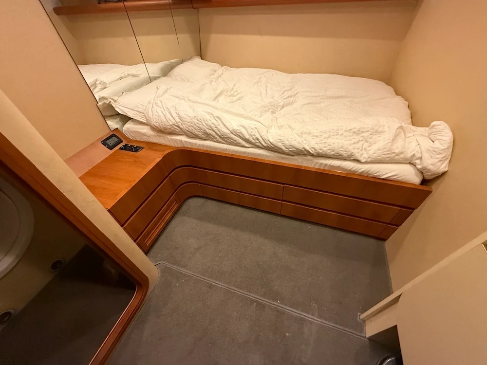 Small hotel room with a bed covered in white bedding, a built-in wooden nightstand, and a mirror behind the bed. The bed is on a platform with drawers beneath, and there's a gray carpeted floor.