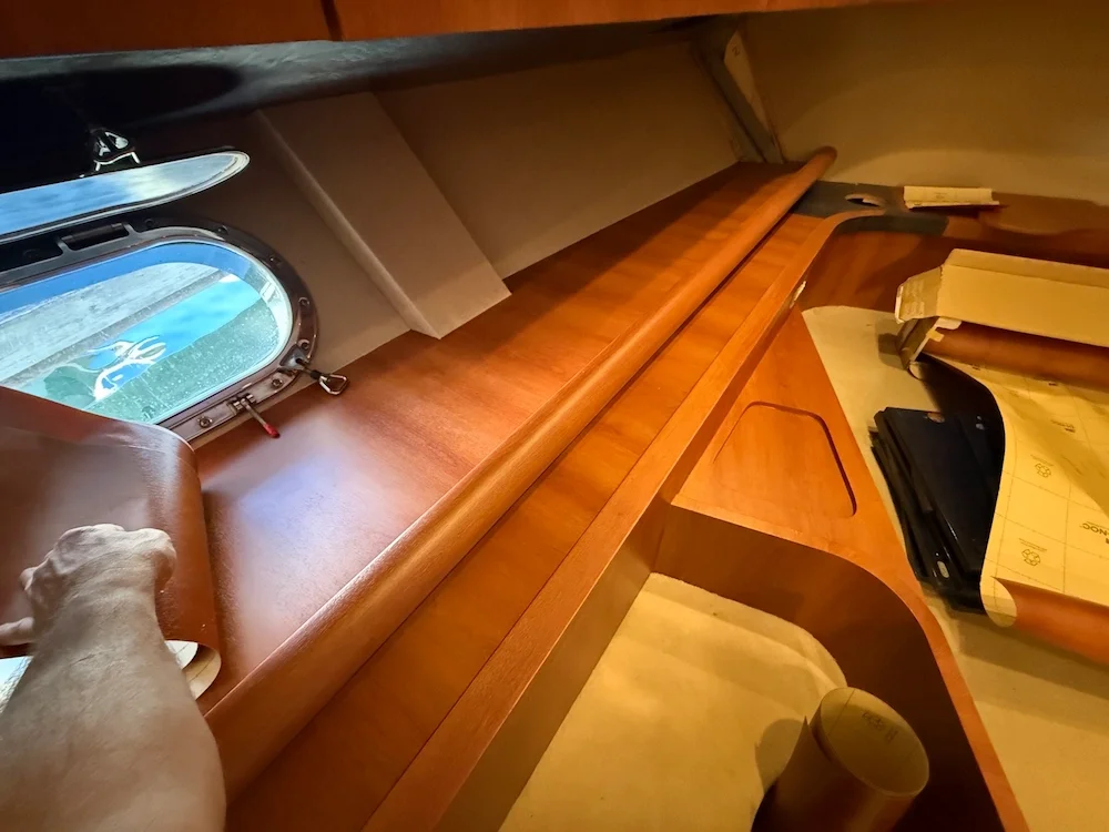 View of the interior of a boat cabin with a wooden desk, small porthole window, and various objects including papers, a folder, and a cup.