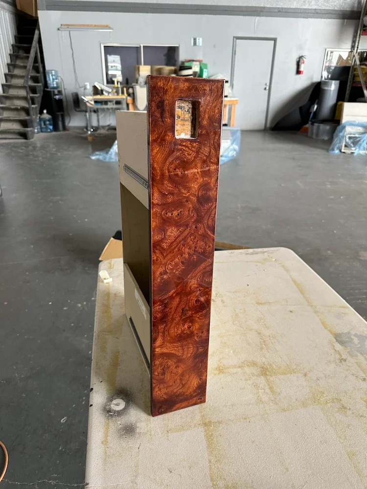 Close-up of a wooden bookend with a glossy, reddish-brown finish, placed on a beige, textured surface in a workshop setting.