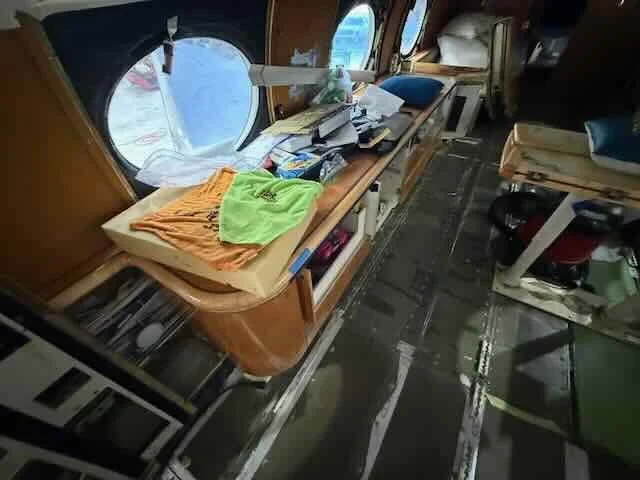 Inside a small airplane cabin, showing a wooden counter with papers, books, and clothes on top. Windows are visible, and the floor appears worn with visible metal strips. There are seats and storage compartments.