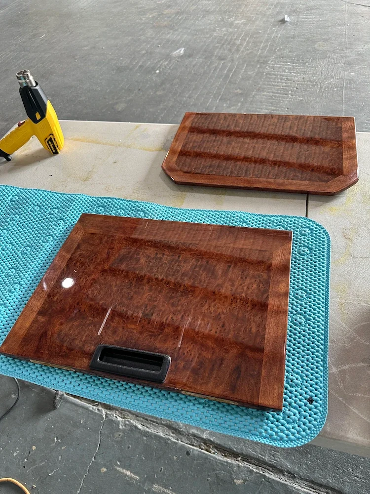 Wooden covers, possibly for furniture or a musical instrument, placed on a blue textured mat on a worktable with a craftsman's glue gun and a piece of white cloth nearby.