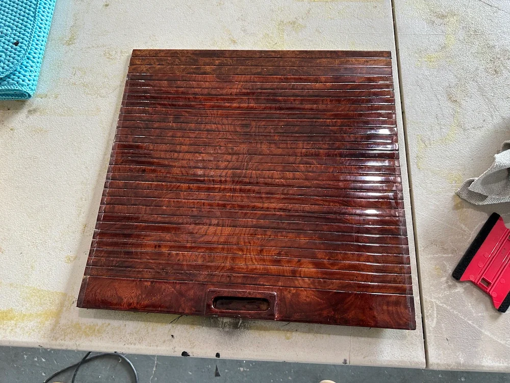 A square wooden panel with horizontal slats resting on a work surface, surrounded by tools and fabrics.