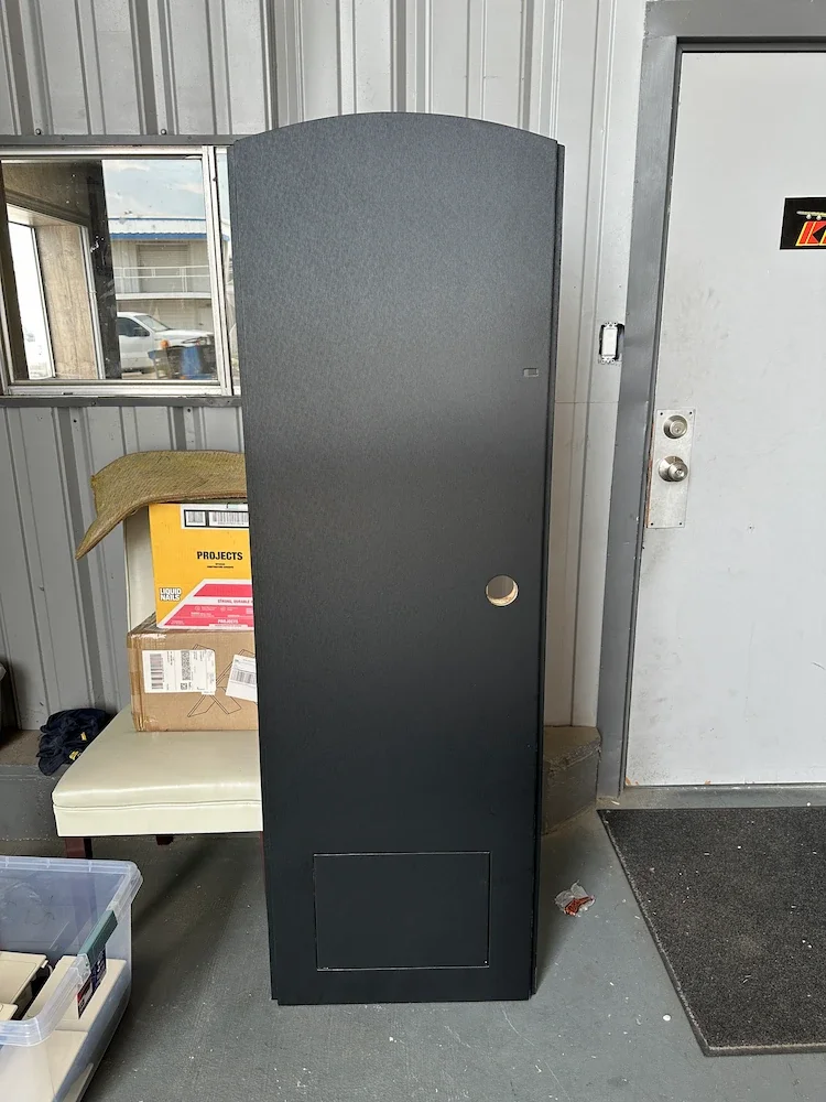 A tall, black storage cabinet with a round hole near the middle, placed against a metal wall inside a building. There is a cardboard box and a cloth on a chair nearby, with a small window and an outdoor scene visible in the background.