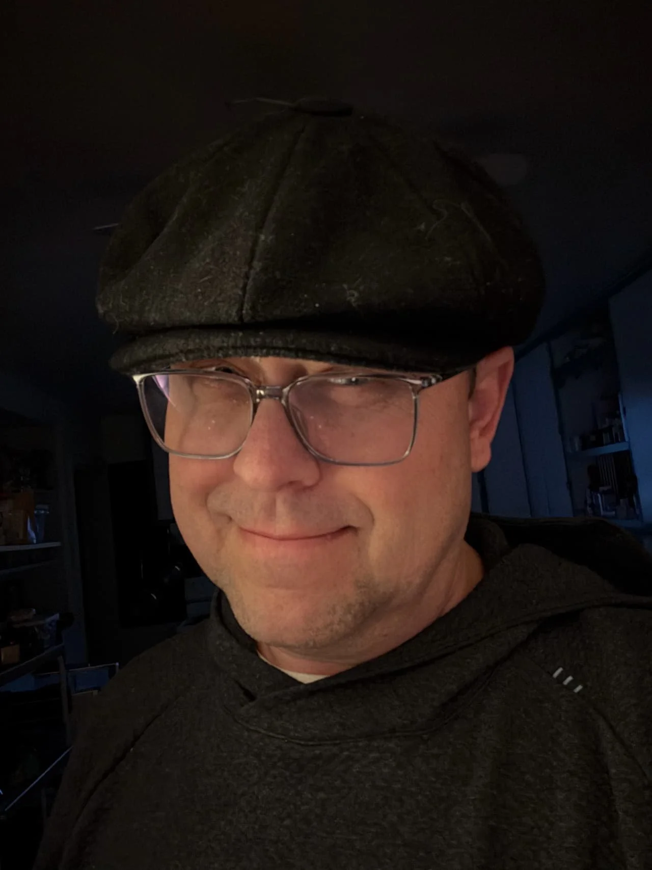 A man with glasses and a dark flat cap smiling, indoor background with shelves and books.