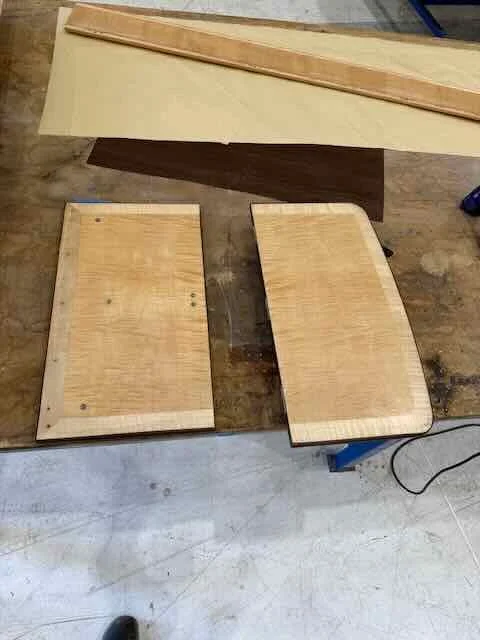 Two unfinished wooden tabletops on a workbench, with a larger piece of wood in the background.