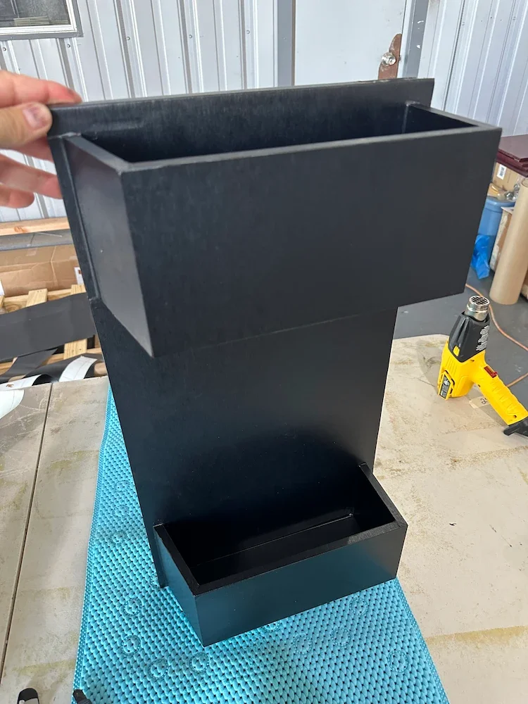 A black wooden planter box in a workshop setting, with a hand holding the top section open. The workspace has a yellow tool, a blue mat, and various supplies in the background.