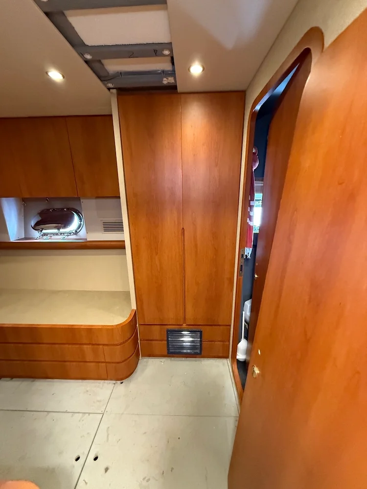 Interior of a small room or cabin with wooden cabinetry, a beige countertop, a small closet with wooden doors, an air vent at the bottom, and part of a doorway leading to another room.