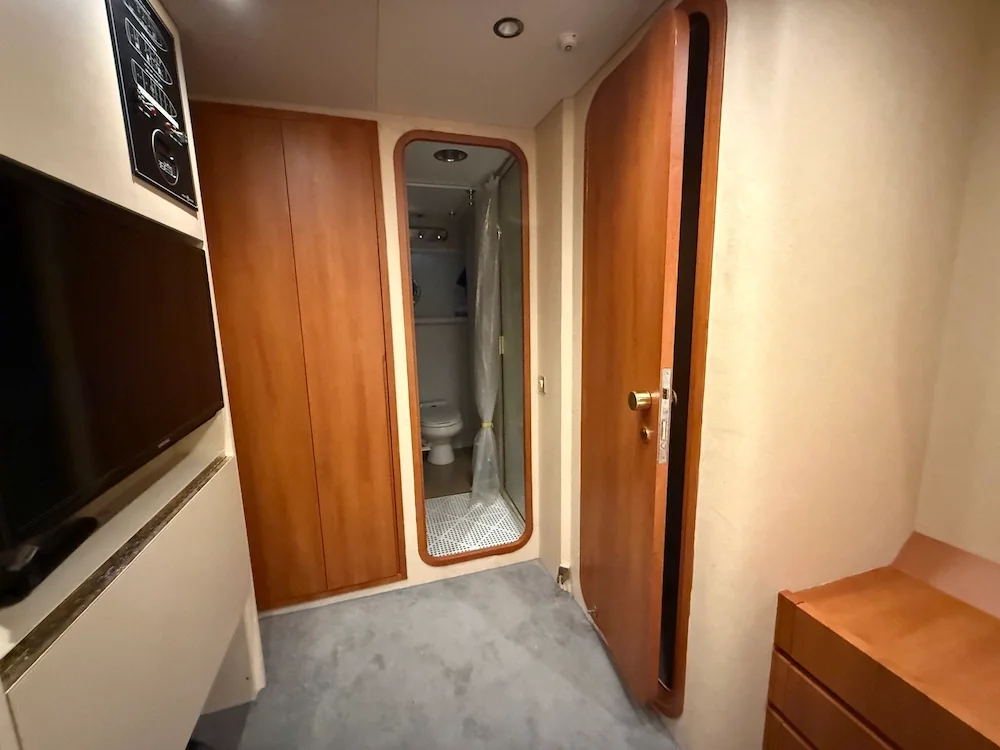 Interior of a small room with wooden doors, a mirror, a TV, and a bathroom visible through an open door, with beige walls and carpeted floor.