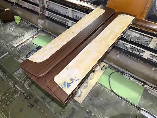 Two wooden tabletops, one dark brown and one light beige with blue paint splatters, resting on a workbench in a workspace with electronic equipment and wiring.