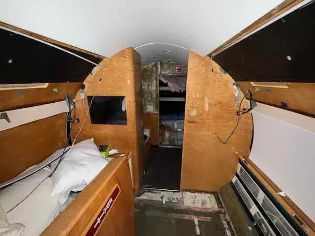 Interior of an airplane in remodelng, with damaged wooden panels, exposed wiring, a bed with pillows, and scattered items.