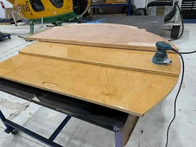 Wooden work table with an electric sander resting on it, in a workshop with tools and equipment in the background.