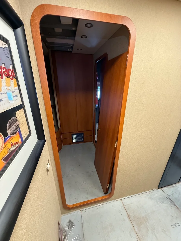 A mirror with a wooden frame mounted on a beige wall, reflecting a hallway with wooden furniture and a partially open wooden door.