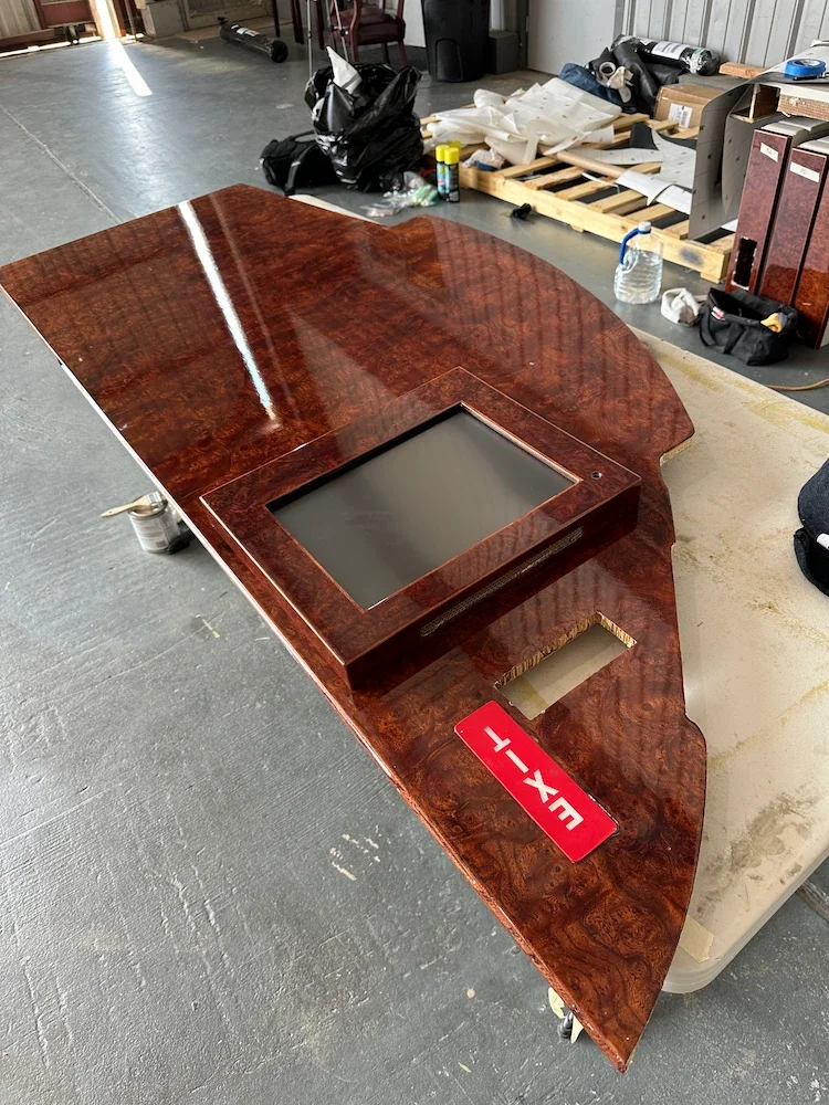 A large, polished wooden conference or executive desk with a built-in monitor or screen area, situated in a workshop or warehouse environment with various tools, supplies, and packaging materials in the background.