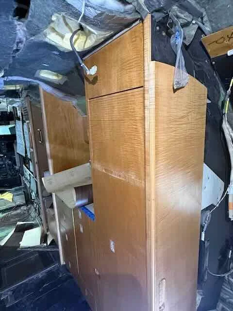 A cluttered room with a wooden cabinet or dresser surrounded by various electronics, cables, and miscellaneous items.