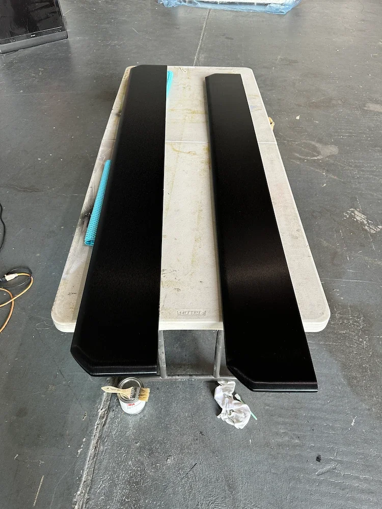 Two black car spoilers placed on a white plastic table in a workshop or garage environment.