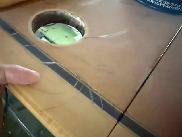 Close-up of a woodworking table with a protruding dowel or bolt, a hand holding the edge of the table, and a hole in the table showing a green object underneath.