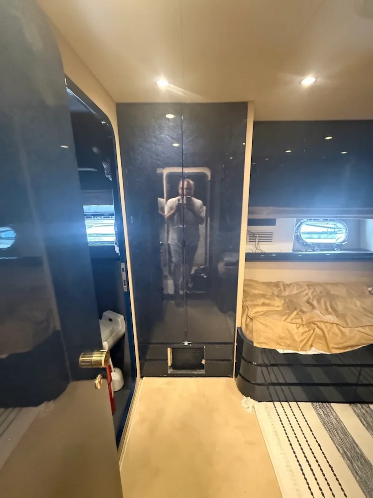 Interior of a yacht or boat cabin with a bed, a porthole, a mirrored wardrobe, and a person taking a photo reflected in the mirror.