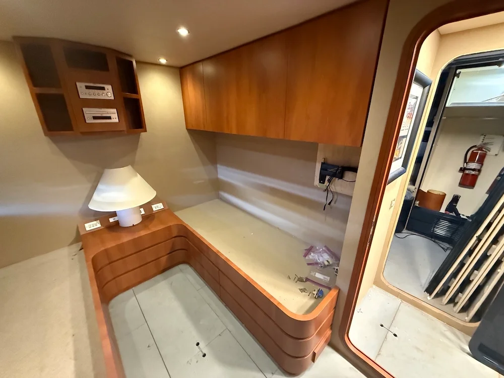 Empty built-in wooden desk with a white lamp, drawers underneath, and wall-mounted cabinets in a room; an open door to an adjoining space with a fire extinguisher visible.