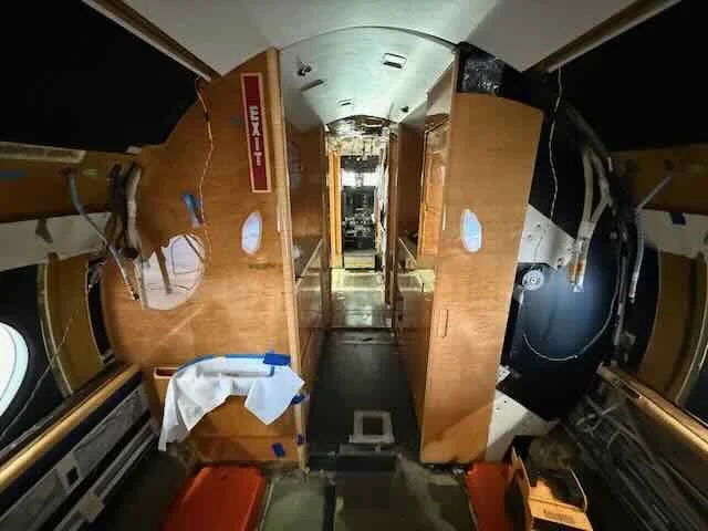 Interior of an aircraft's cabin with wooden panels, small windows, an exit sign, and some equipment including a white cloth and orange containers on the floor.
