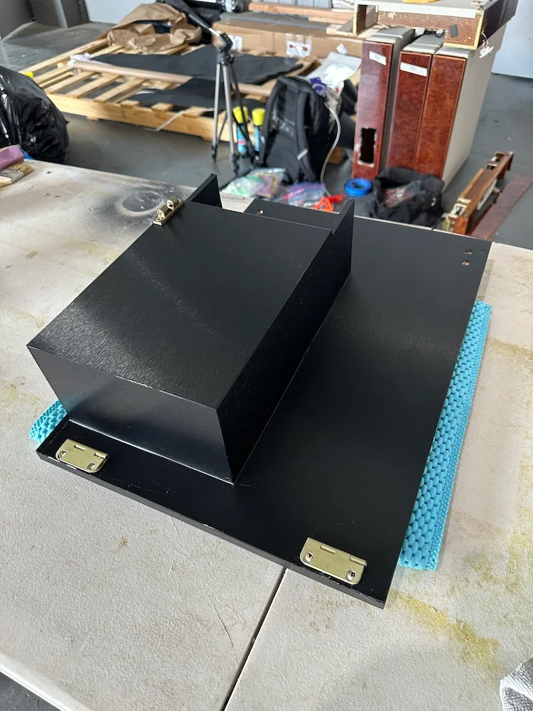 Black electronic device with metal hinges placed on a table in a cluttered workshop or garage.