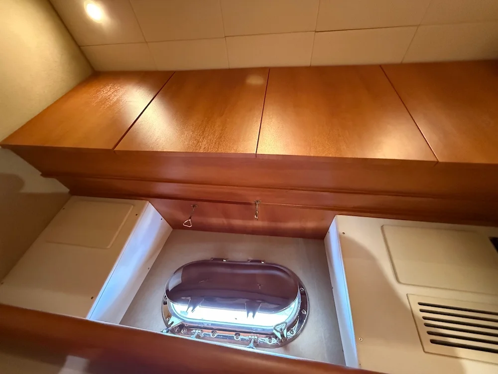 Interior view of a section on a boat, showing a wooden counter with closed cabinets above and a porthole window below, with a tile wall in the background.