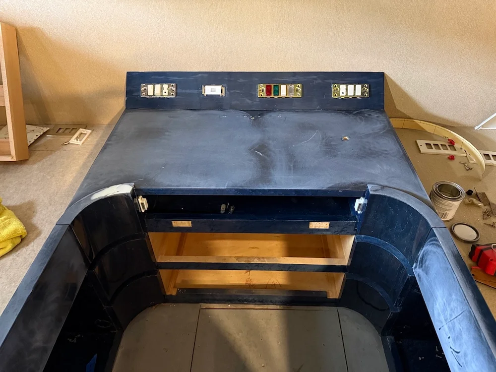 Empty black wooden desk or countertop with an open storage area underneath, situated against a beige wall, with various tools and objects around it.