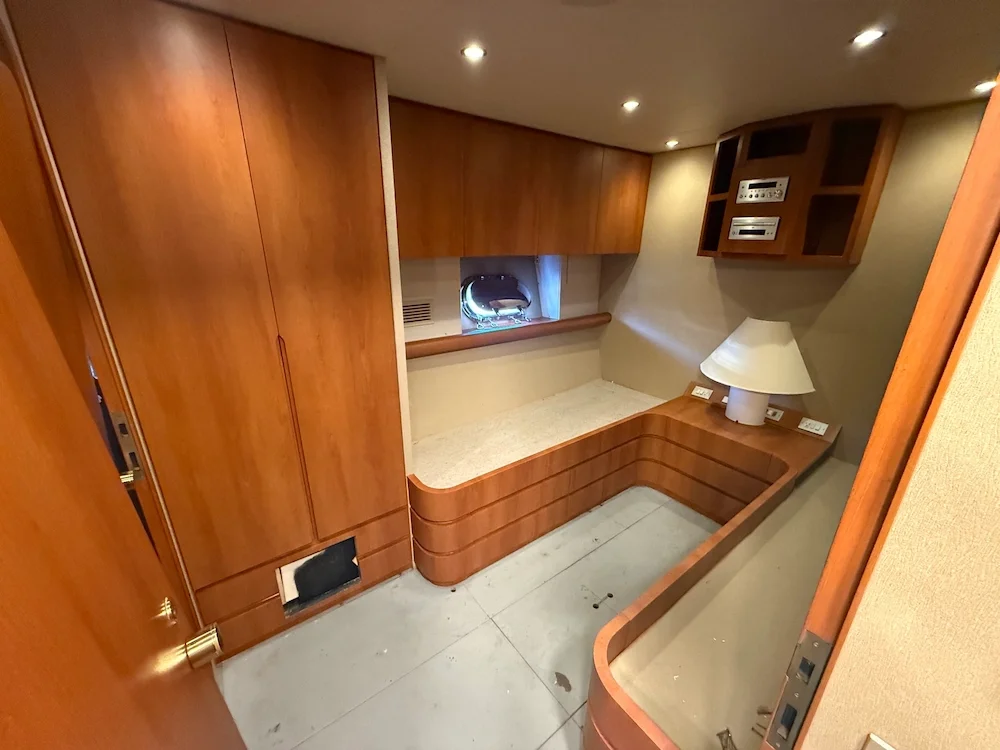 Small room with wooden built-in furniture, a low wooden bed frame, a side table with a white lamp, and a small built-in shelf with electronic equipment. The room has beige walls, wood paneling, and a small window.