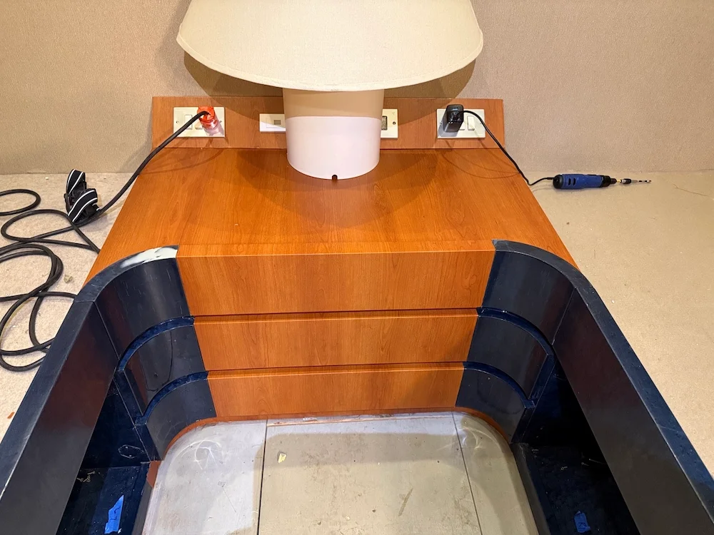 A wooden nightstand with three drawers, a lamp, and charging ports on top, is placed against a beige wall. An electric drill and tangled cords are visible nearby.