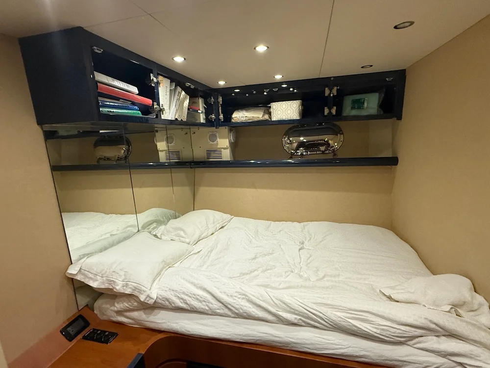 A bed with white rumpled sheets and pillows, underneath a mirrored wall and above a shelf with books, decorative objects, and a tissue box in a room with beige walls and ceiling.