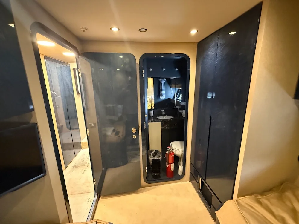 A small bathroom with a black storage cabinet, a mirror, and a partial view of a bed with beige bedding.