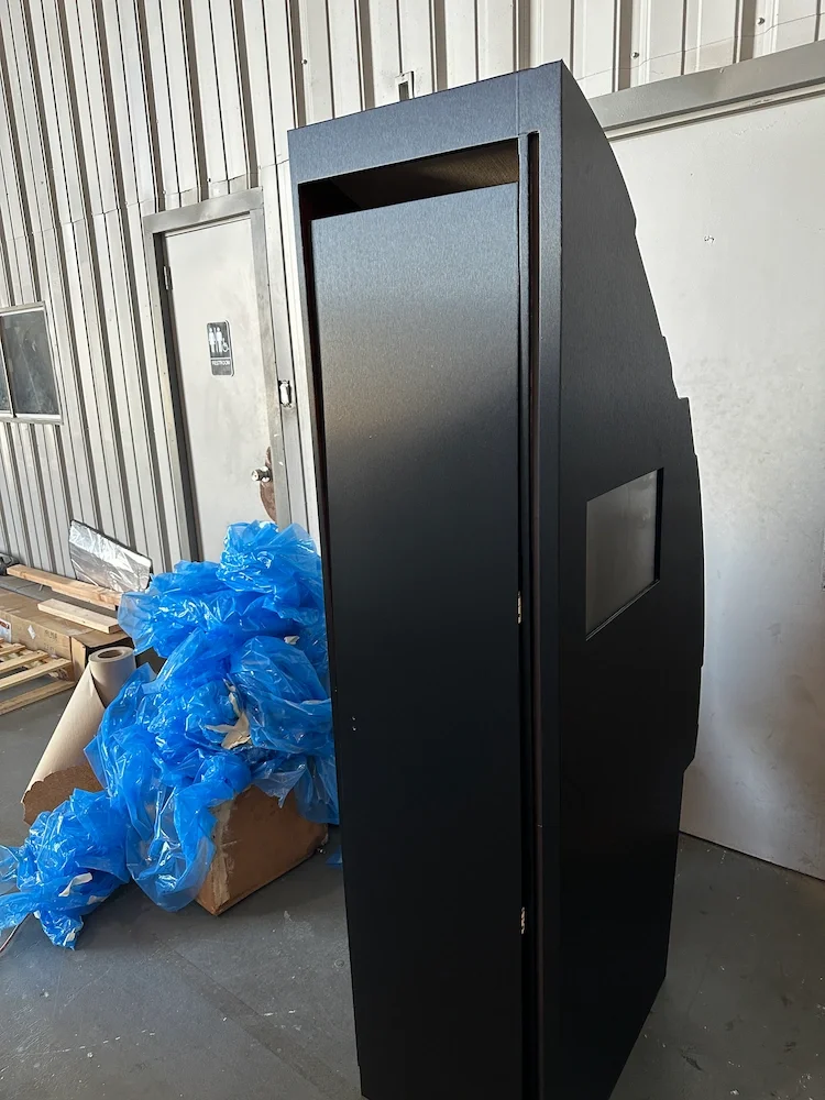 A black kiosk with a small screen on the side in an industrial warehouse setting, next to a cardboard box with blue plastic bags.