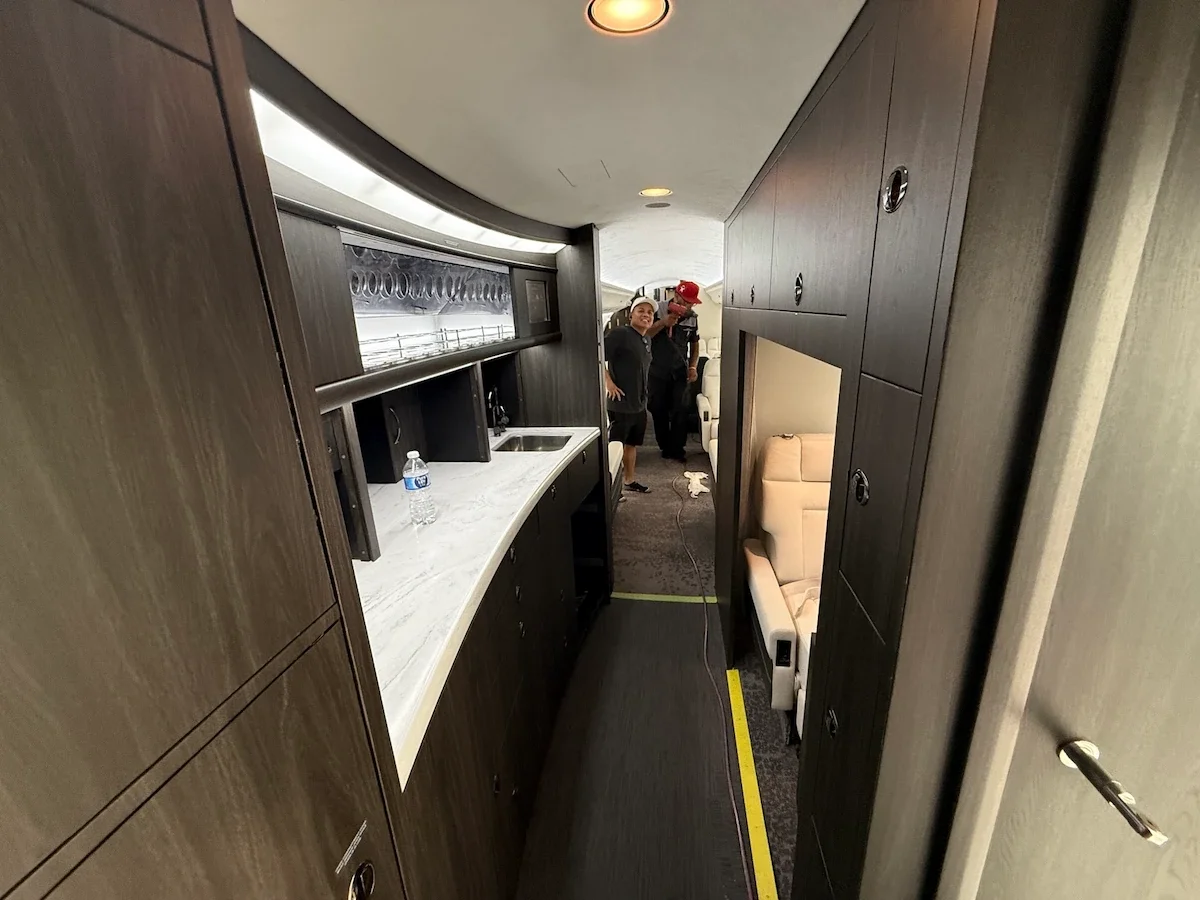 Interior of a private jet or airplane cabin with wooden cabinetry, a small sink, and a water bottle on the counter. Two people are standing in the background near the seating area, one wearing a hat and the other wearing a red cap. The photo is taken from a narrow corridor.