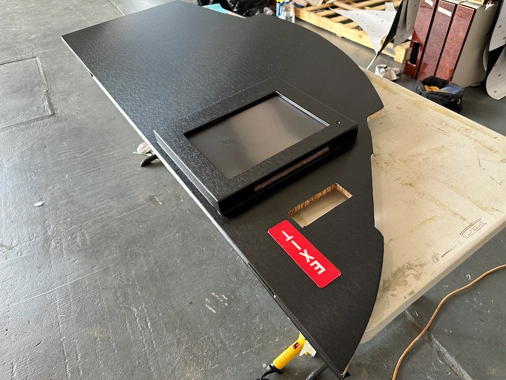 A flat black dashboard with a built-in digital display, labeled 'EXX-L', resting on a white worktable in a workshop or warehouse setting.