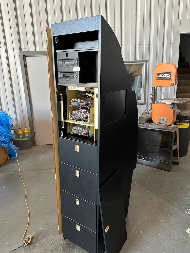 A tall black arcade-style cabinet with the back panel removed, exposing internal electronic components, situated in a workshop or garage.