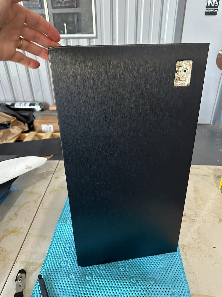 A person holding a black rectangular panel with a small electrical outlet on the upper right corner. The panel is on a blue mat on a workbench, with a pen nearby and storage items in the background.