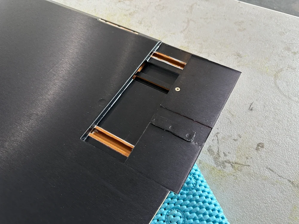 Close-up of a black electronic device with a partially open panel, revealing copper wiring inside, on a gray surface with a blue textured mat nearby.