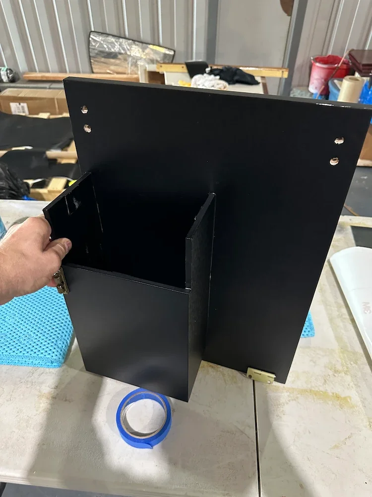 Person opening a small black cabinet or box on a worktable in a workshop setting.