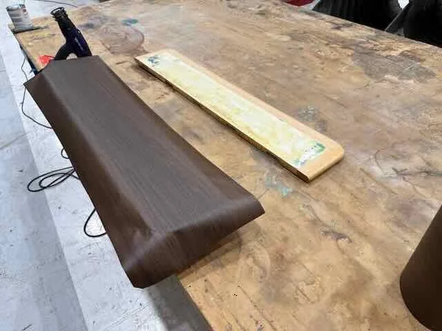 A piece of dark brown wood being laminated or coated, resting on a cluttered wooden workbench with a yellowish wooden board and a glue bottle nearby.