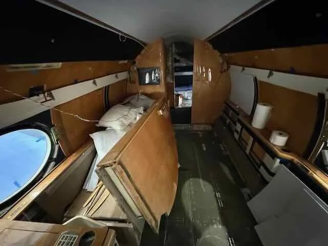 Interior of an airplane with wooden paneling, showing seats and storage compartments, with some items like pillows, toilet paper rolls, and equipment scattered inside.