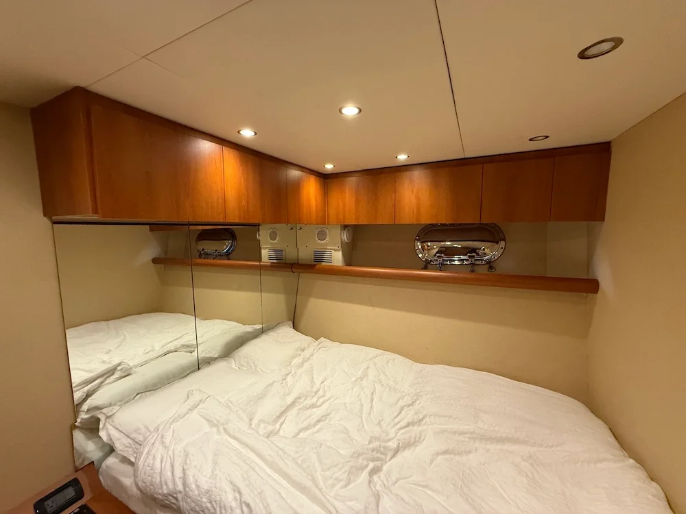 A cozy bedroom with a bed, mirrored closet doors, wooden cabinets above, and a shelf with nautical decorations. Recessed ceiling lights illuminate the space.