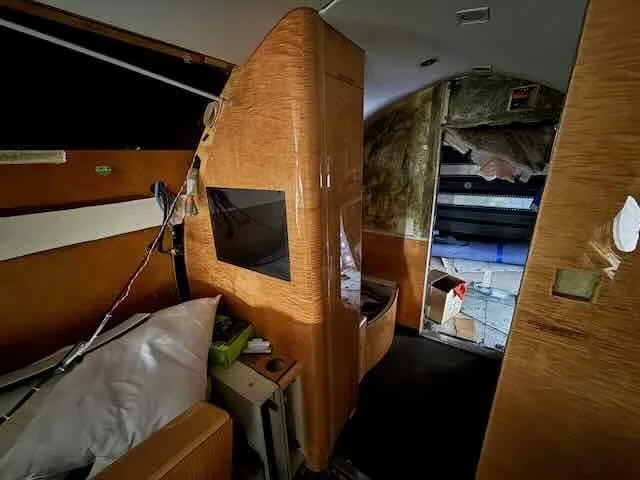 Inside a small airplane cabin, showing a bed with pillow, a small TV mounted on a wooden partition, and a narrow hallway with a bathroom door open to the right.