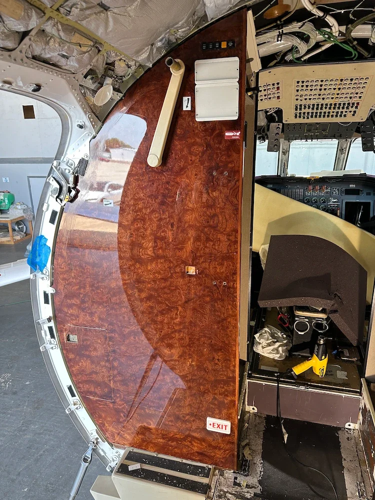 Interior of an aircraft cockpit with a large, polished wood panel partially installed, various control panels, wiring, and tools visible, including a yellow drill and a piece of foam padding.