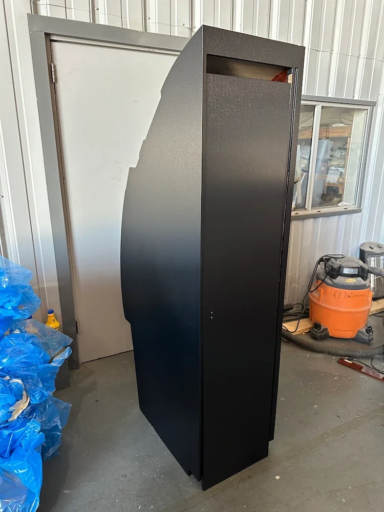 A tall black vending machine or electronic kiosk in a warehouse or storage area with a door and window in the background, and orange and blue storage bins and equipment on the floor.