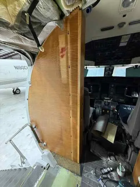 The cockpit of an airplane with a large wooden partition. The aircraft is on the ground, with part of the runway and another aircraft visible outside. Inside the cockpit, there are various controls, screens, and equipment.