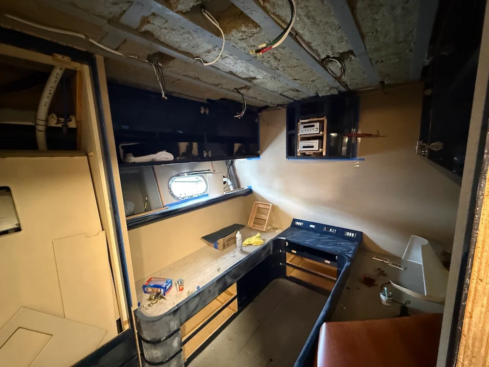Construction or renovation site showing a partially installed countertop, exposed insulation and electrical wiring on the ceiling, and various tools and materials on the surface.